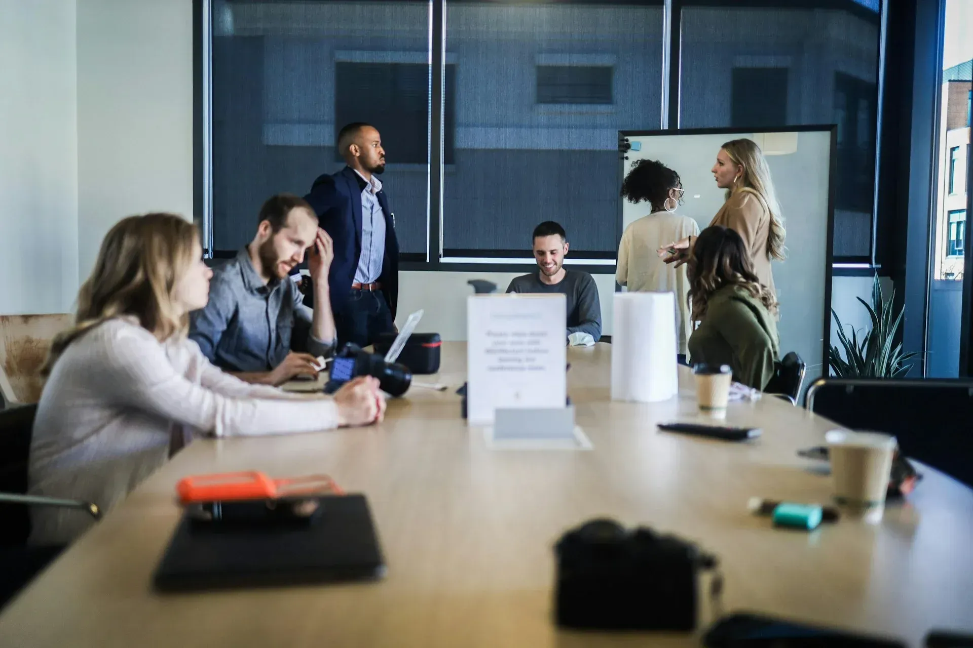 People in a meeting room, some seated around a table, others standing near a whiteboard.