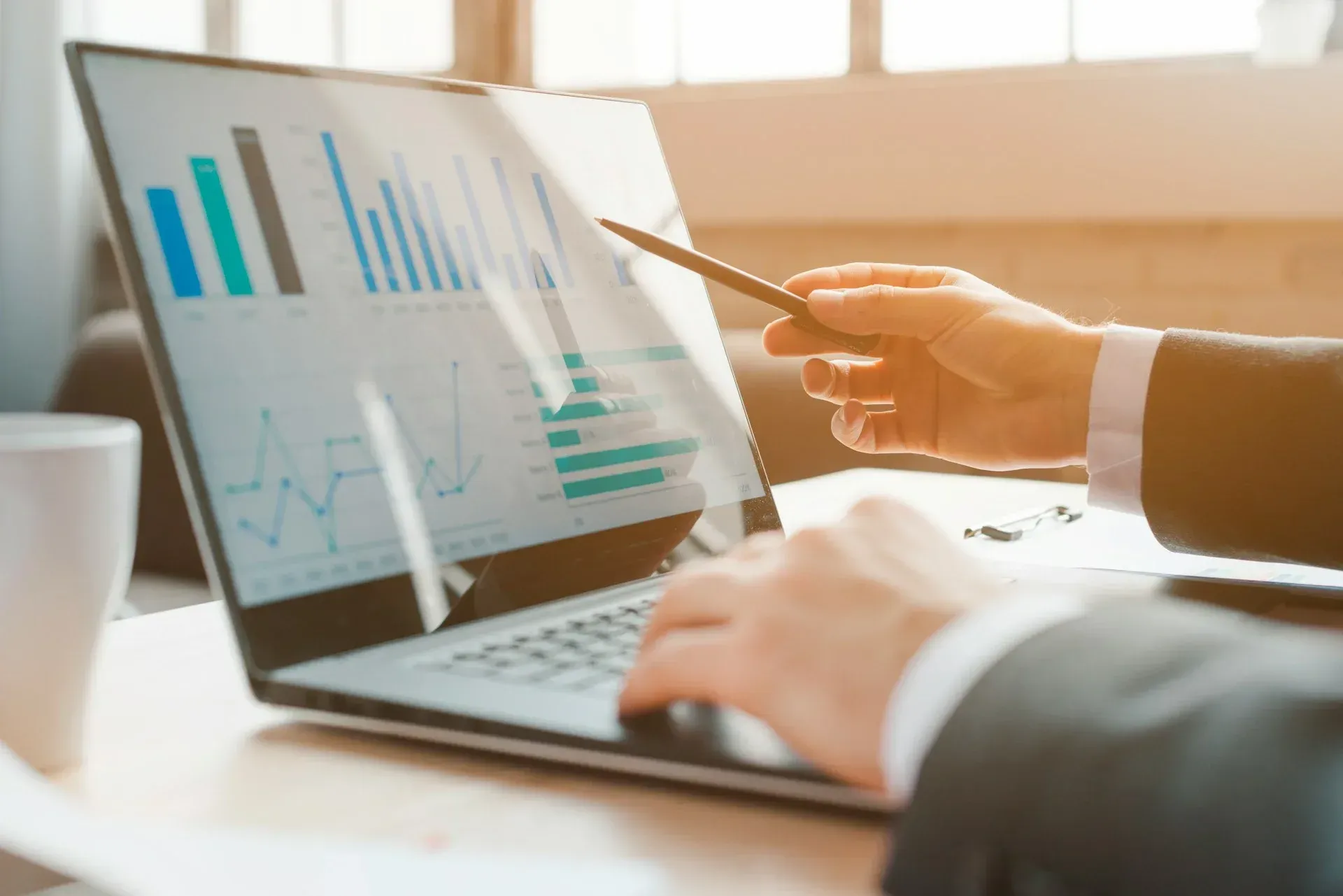 Person in suit pointing at a laptop screen with financial charts. Sunlight streams in.
