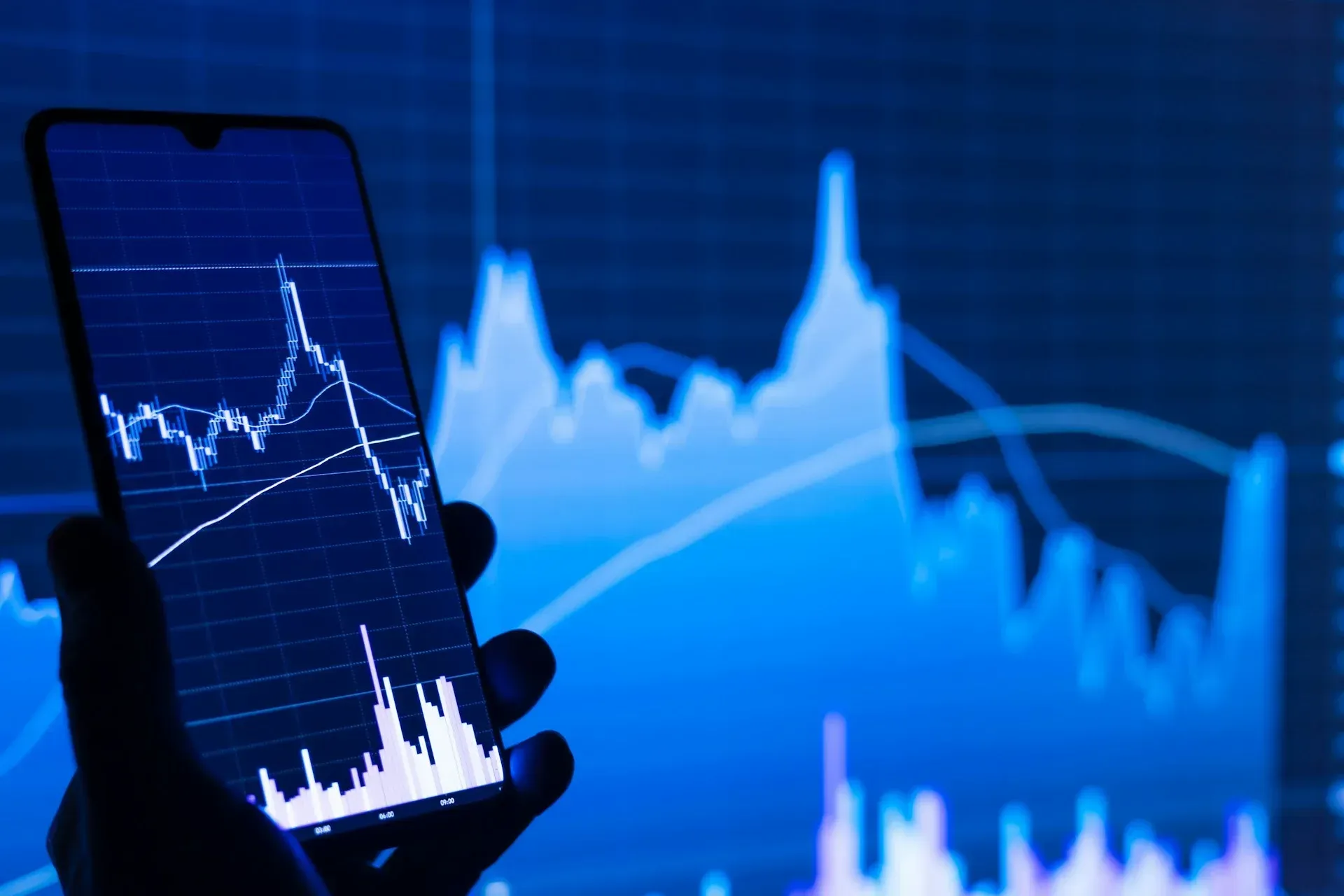 Hand holding smartphone displaying a stock market graph, overlaid on a larger blue financial chart.
