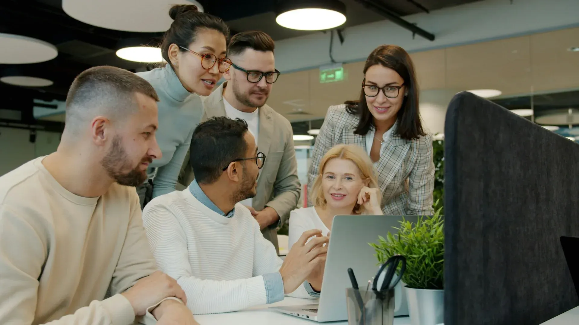Group of people collaborating around a laptop in an office setting, looking at the screen with expressions of focus.