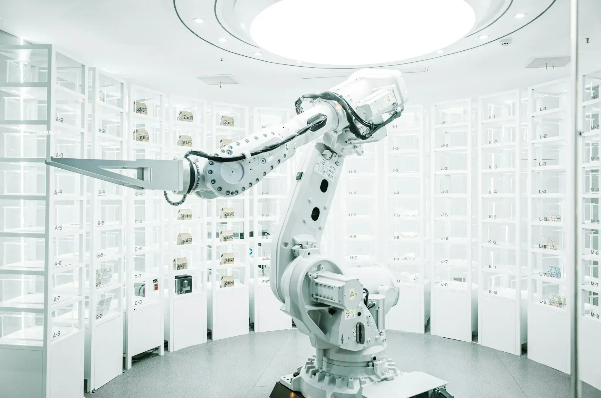 White robotic arm in a circular white room with shelves filled with objects, under a round ceiling light.