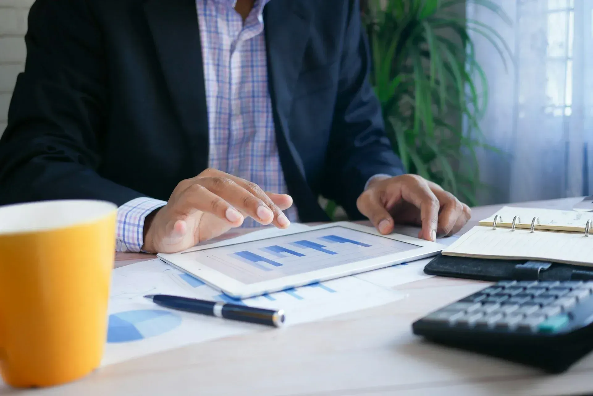 Person in suit analyzes charts on a tablet at a desk with a coffee mug, pen, notebook, and calculator.