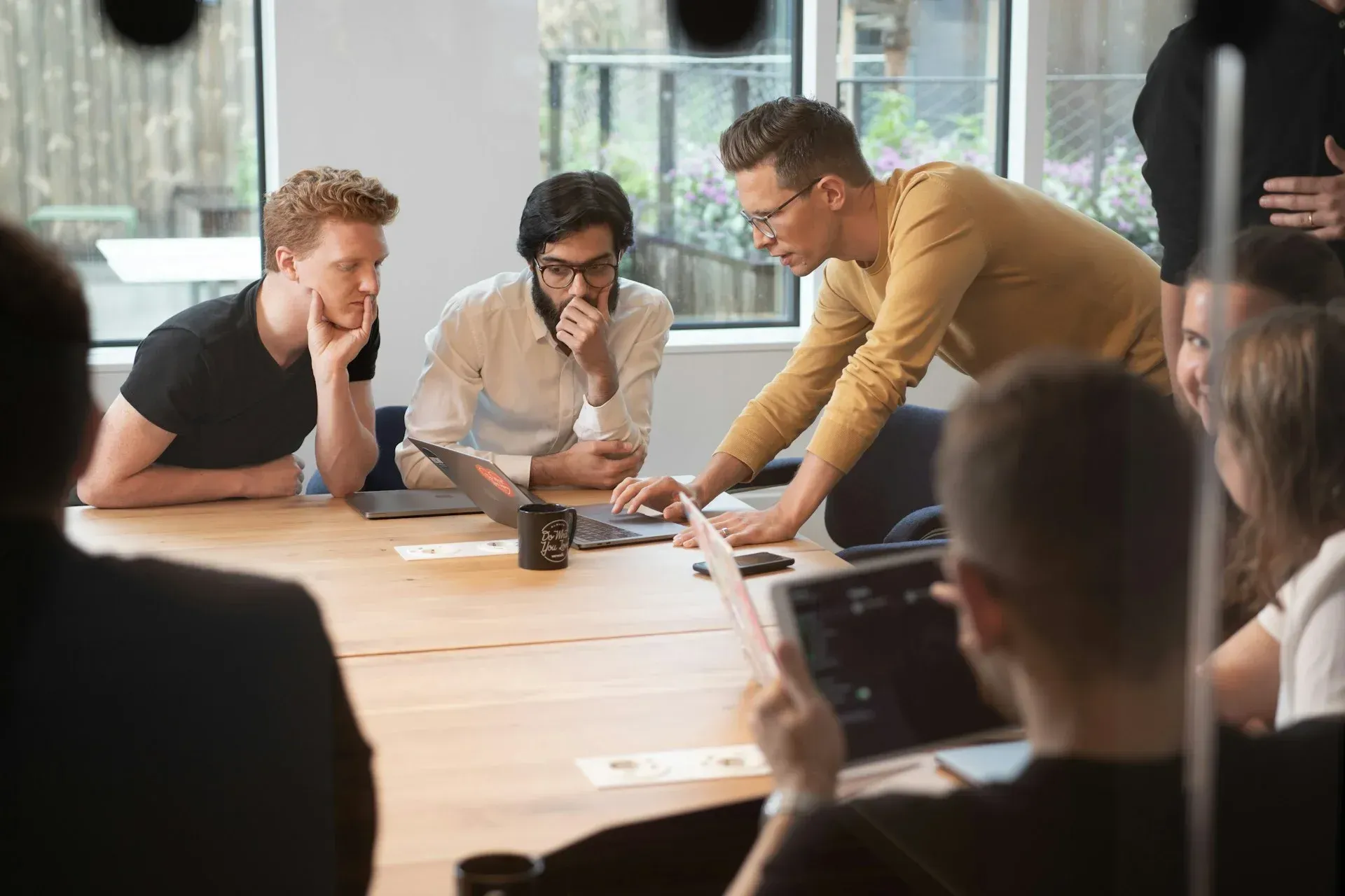 Group of people in a meeting, gathered around a table with laptops, discussing a project.