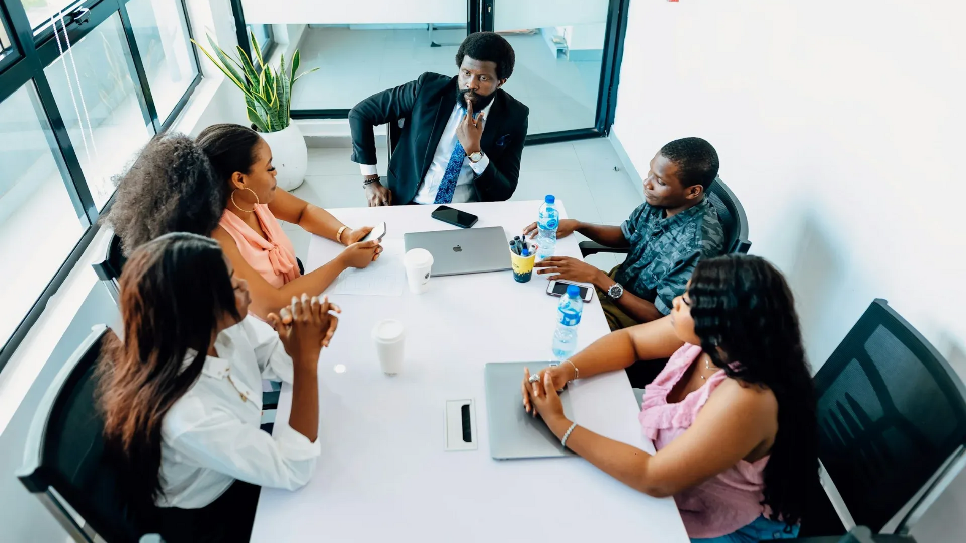 Group of people in a meeting at a white table, one man standing, some with laptops, water bottles, and a plant.