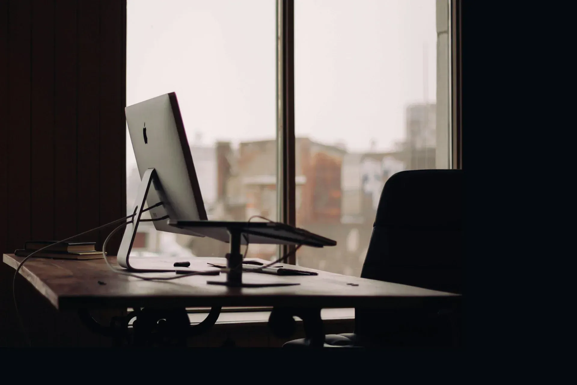 Desk with computer near a window, silhouetted. Outside, brick buildings visible through the glass.