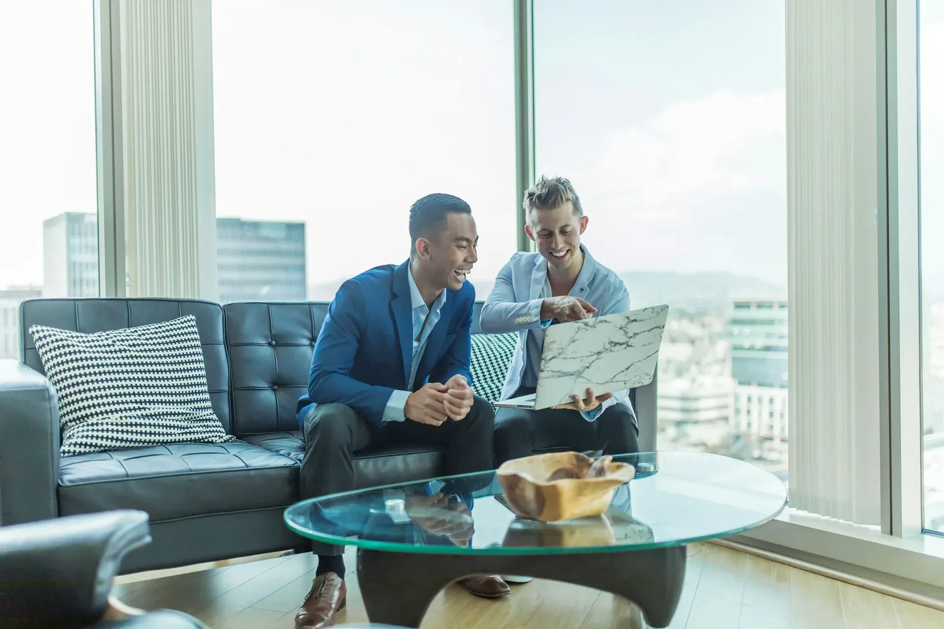 Two people in business attire look at a laptop in a modern room with city views.