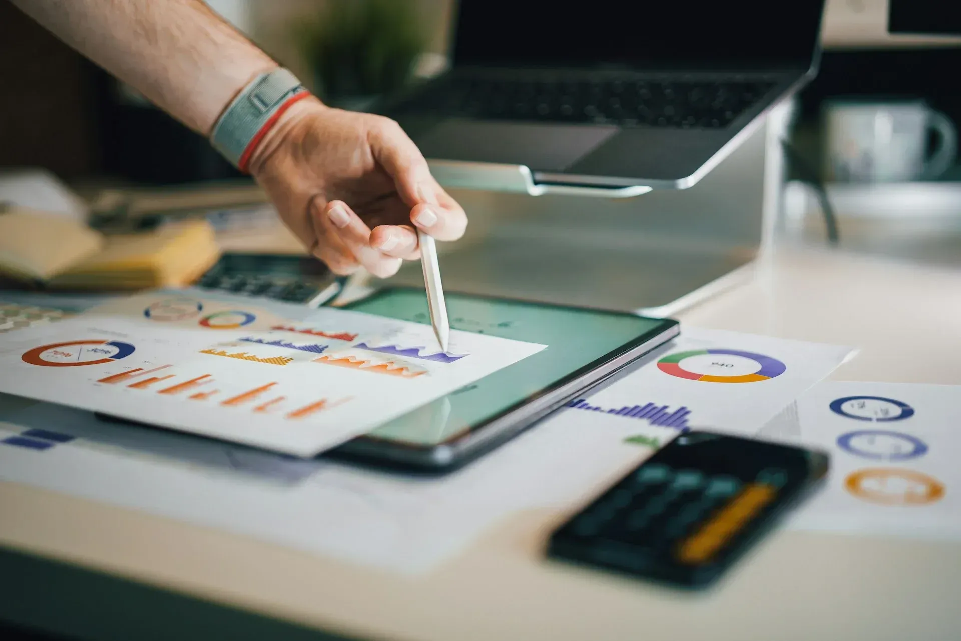 Person pointing at charts on a tablet, with laptop and calculator on a desk.