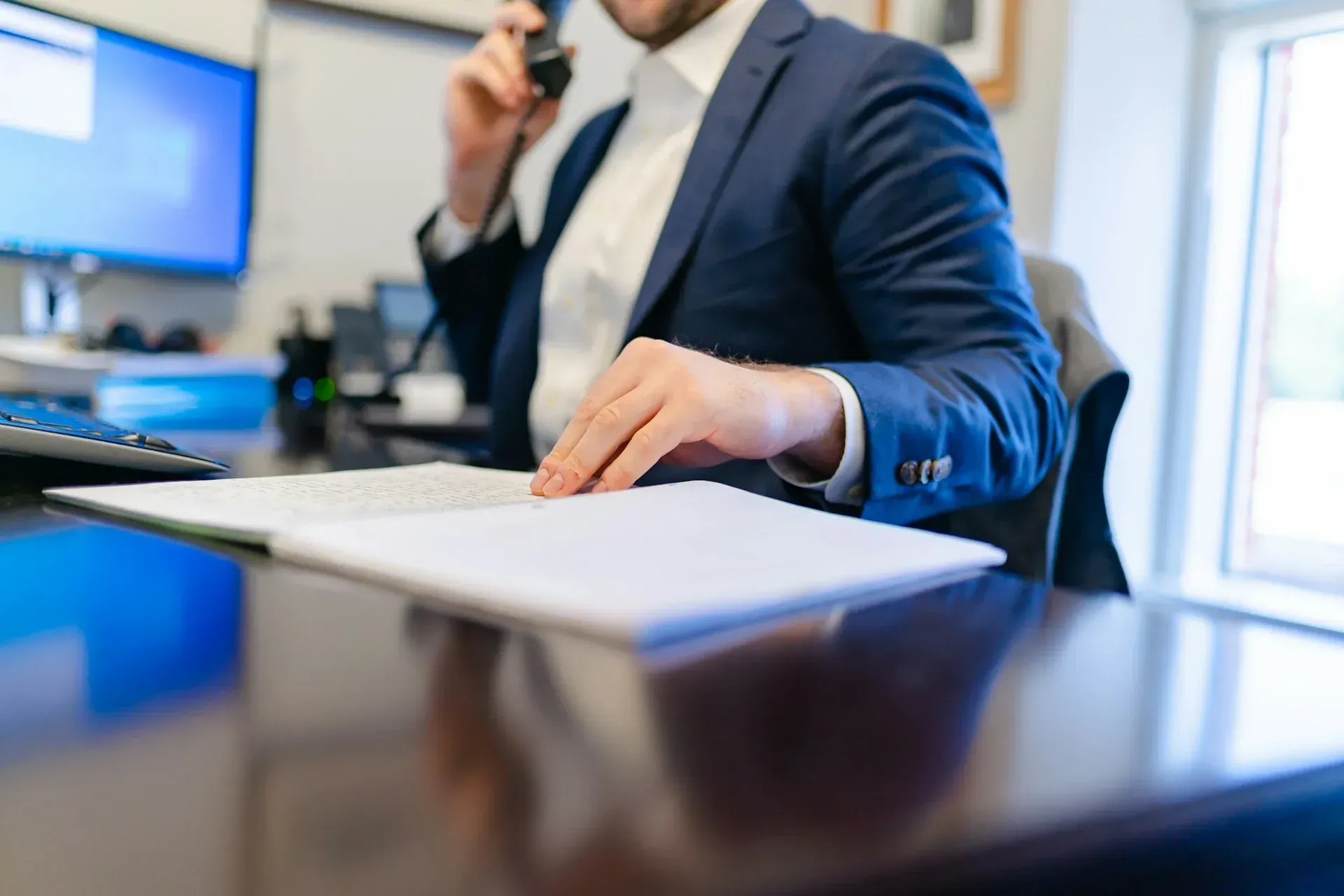 Man in blue blazer on phone, looking at documents at desk. Office setting.