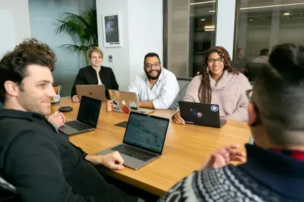 People at a table with laptops in a meeting. Smiling, in an office with a plant.