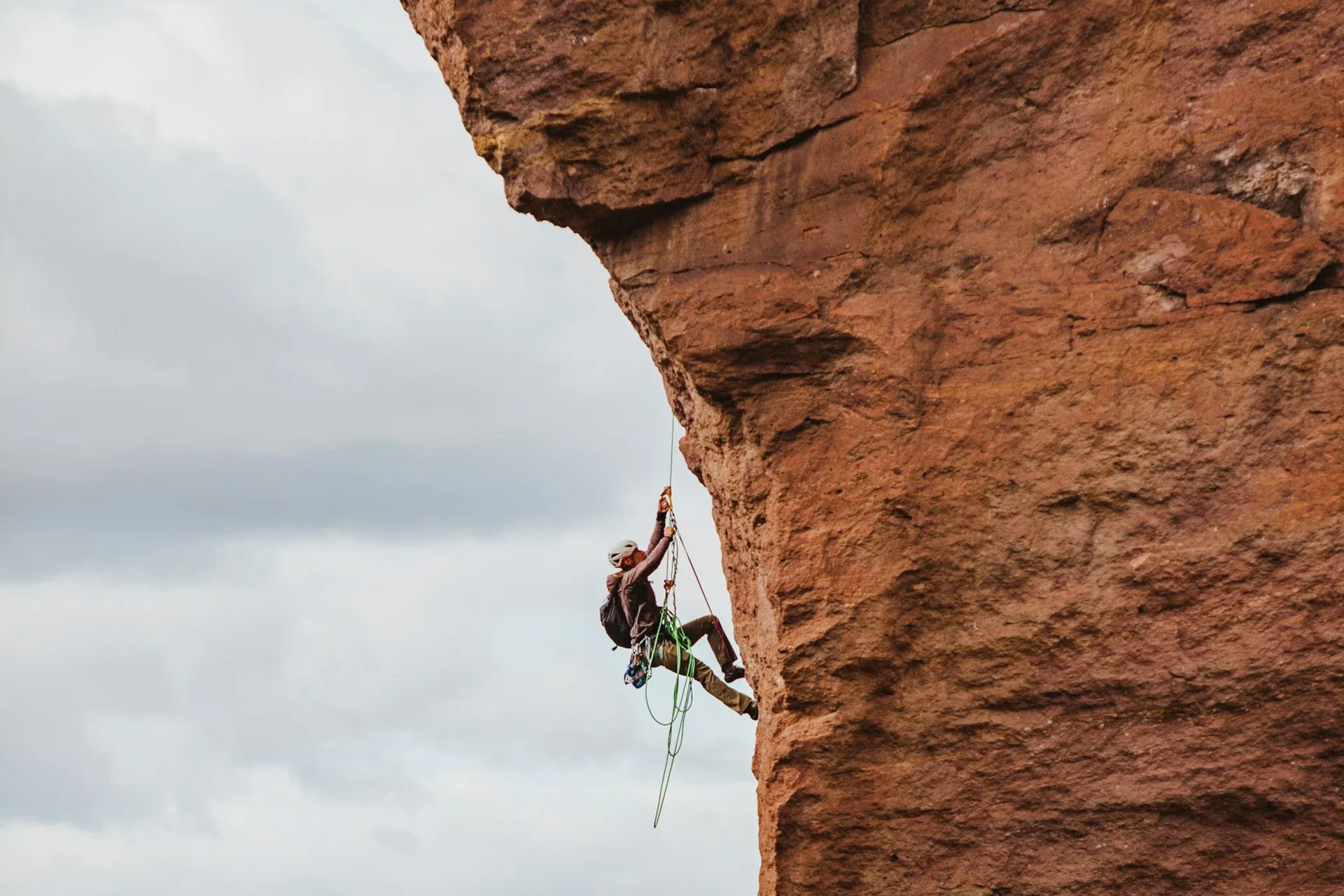 Rock climber on a red rock face, using ropes and gear against a cloudy sky.