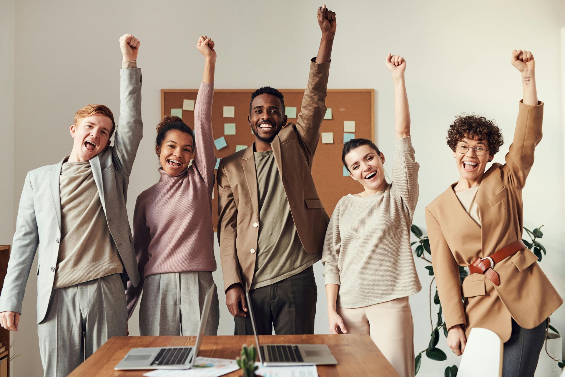 Five people in office, arms raised in celebration; wood desk, corkboard, bright, happy expressions.