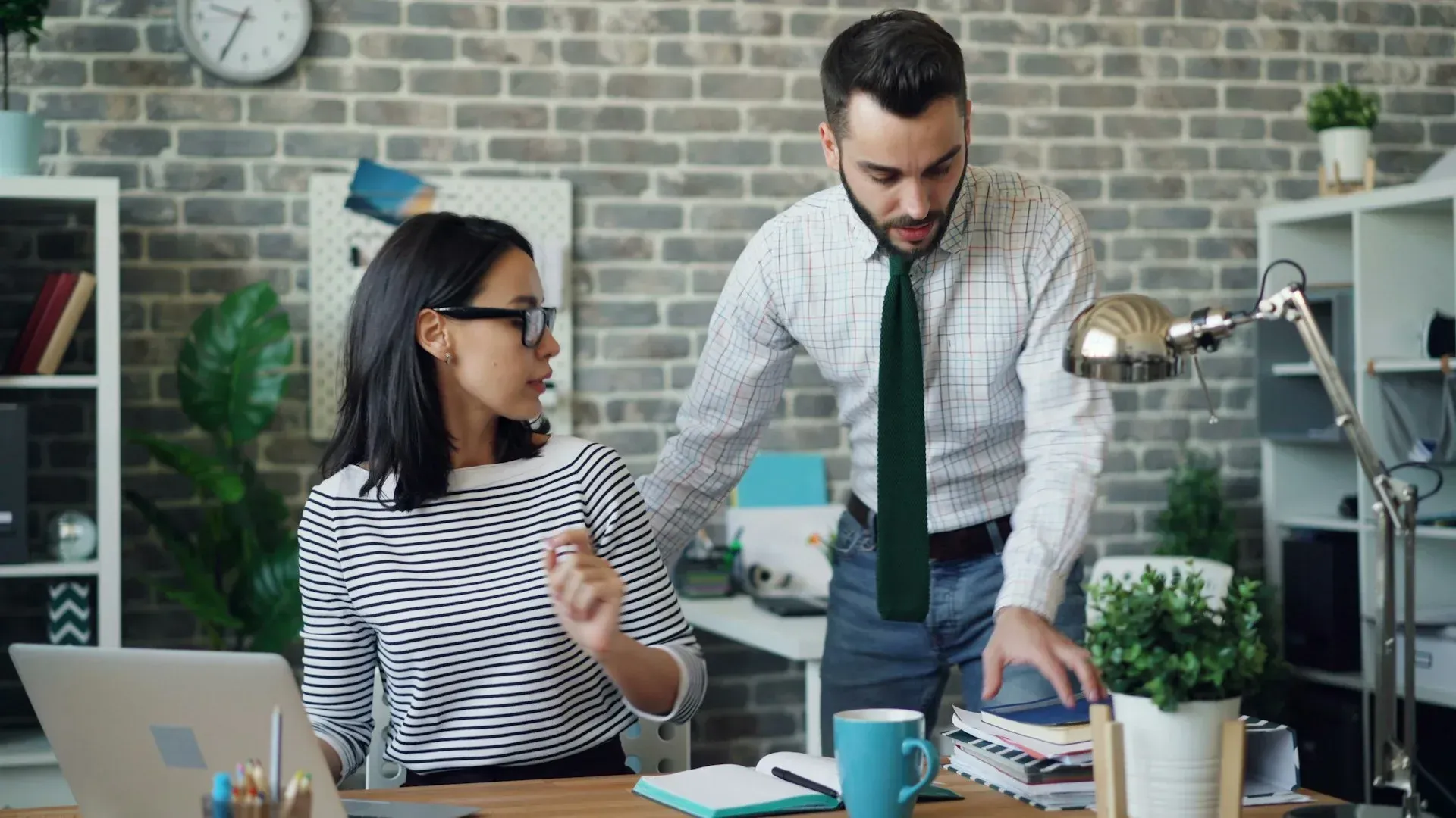 Woman at laptop, man standing over her shoulder looking at paperwork in an office setting.