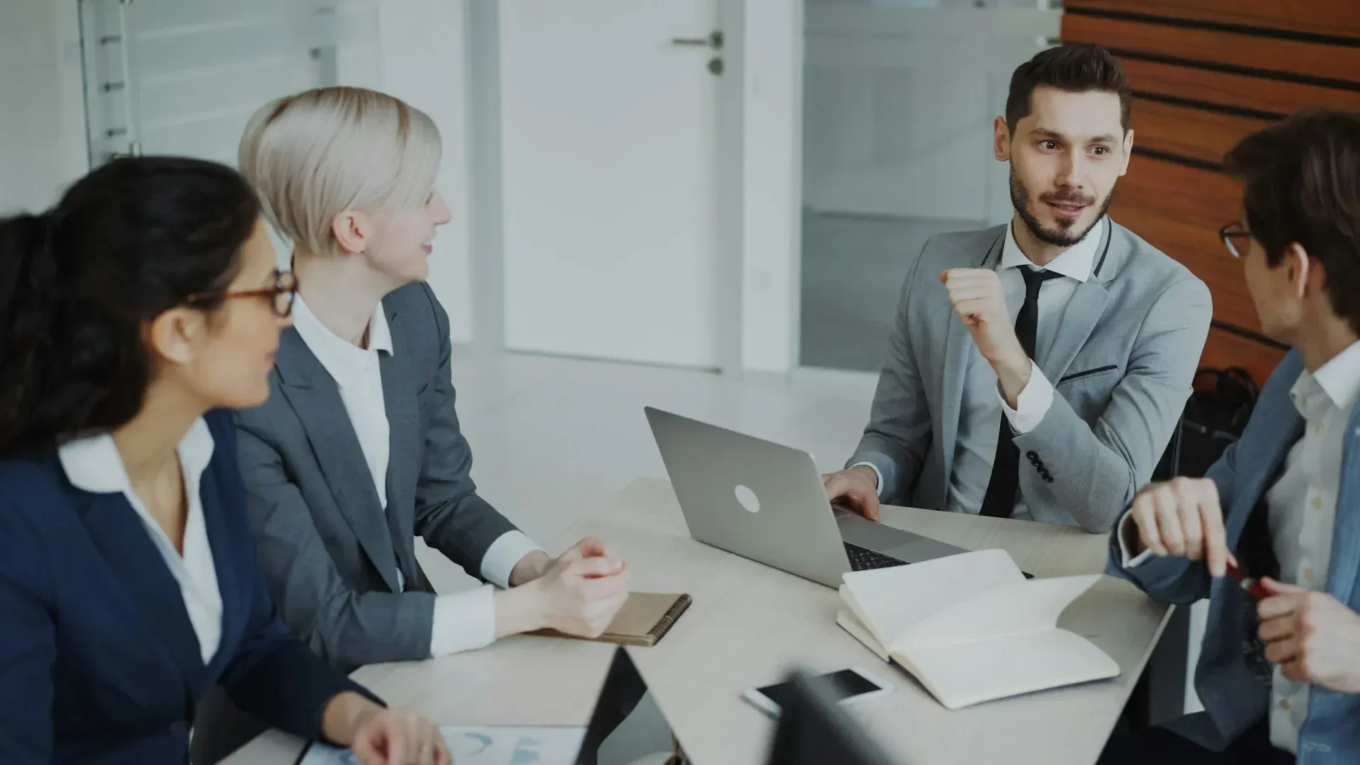 Business meeting: Four people in suits at a table. Man with beard gestures. Laptop and open notebooks present.