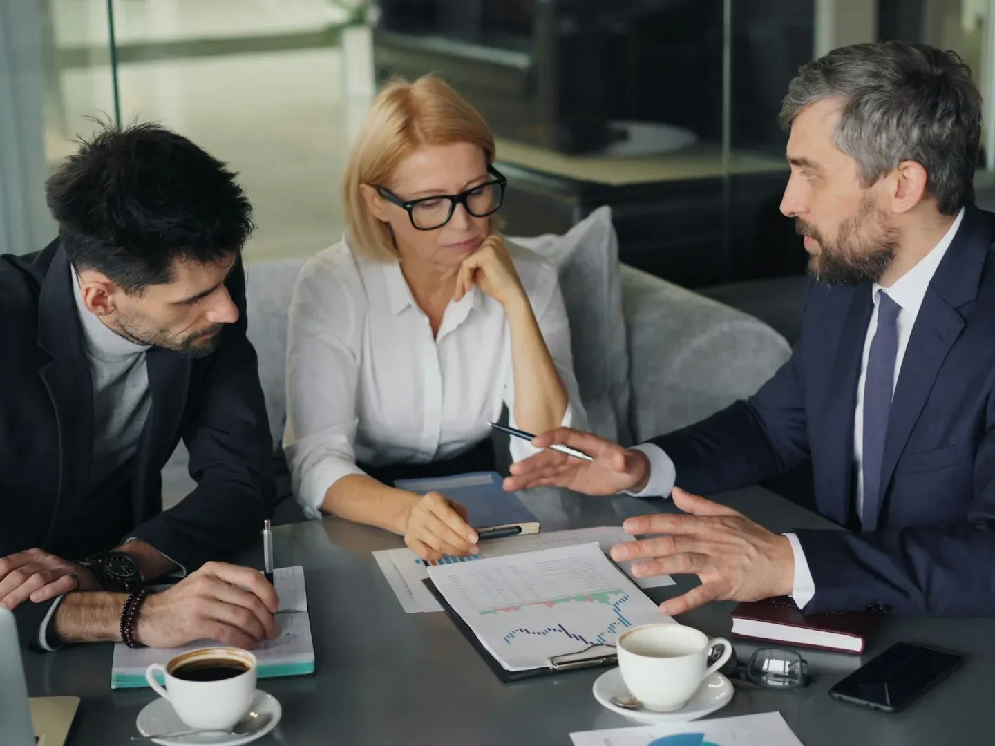 Three people in suits at a table reviewing documents, a woman in glasses listens, a man gestures.